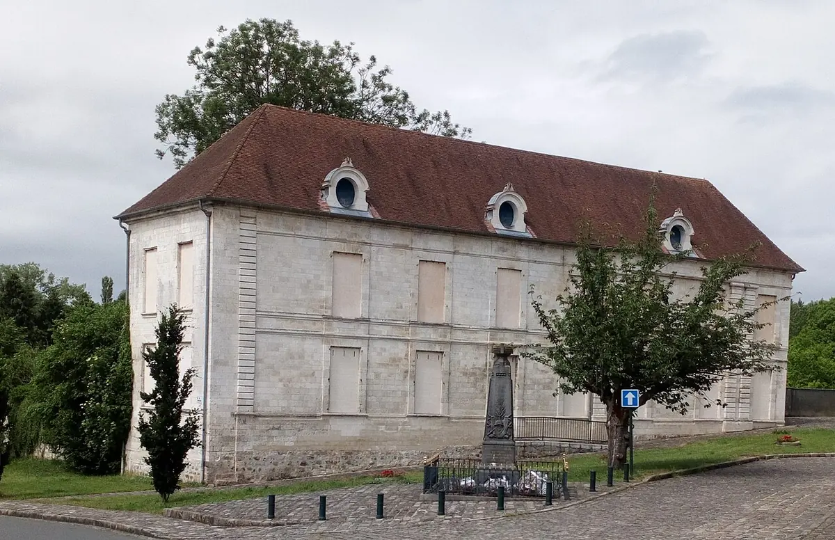 A long two-story stone building with a steep tiled roof and dormer windows beside a small village square