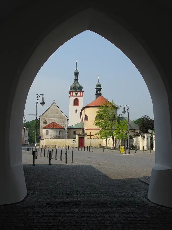 A white church with a tall bell tower and onion-domed towers, seen through a Gothic stone archway across a cobbled square