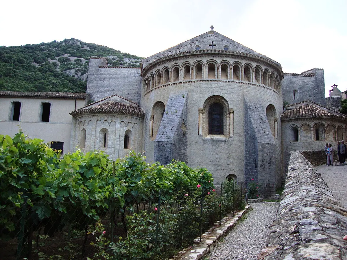 The rounded Romanesque stone apse of an abbey church with a row of small arches under its roof, above a garden of vines and roses