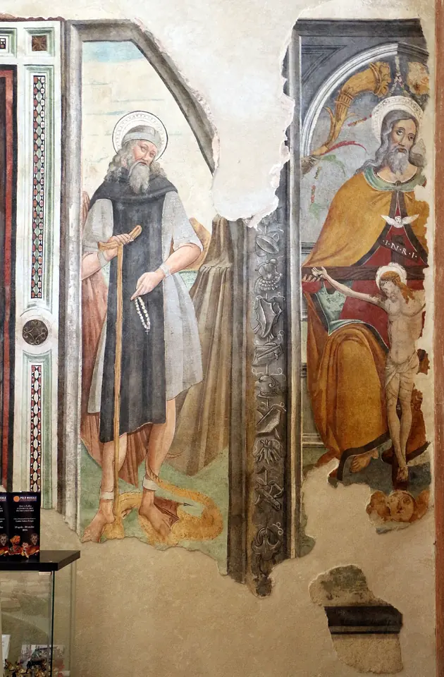 Faded wall fresco of a bearded hermit saint with a halo and a dark habit holding a rosary and staff, painted beside an image of God the Father holding the crucified Christ