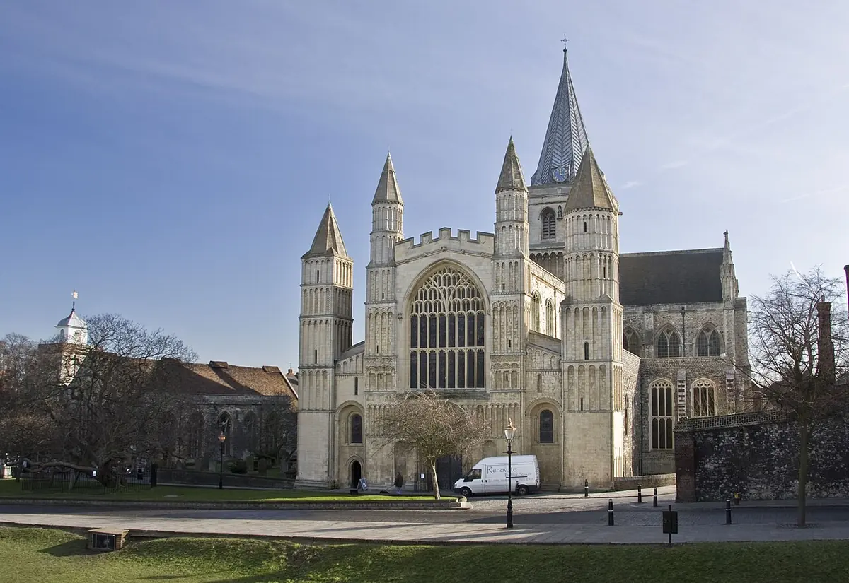 The pale stone west front of Rochester Cathedral with its towers and a large window, under a blue sky, with a lawn in front