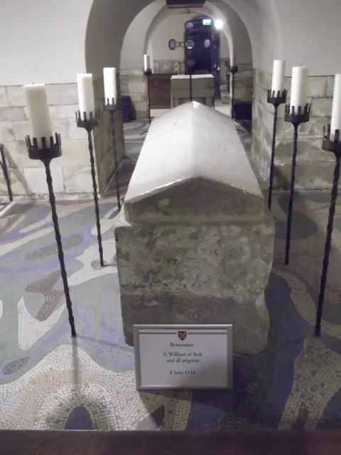 A plain stone coffin with a pitched lid in a white-vaulted crypt, surrounded by tall black iron candlesticks with white candles