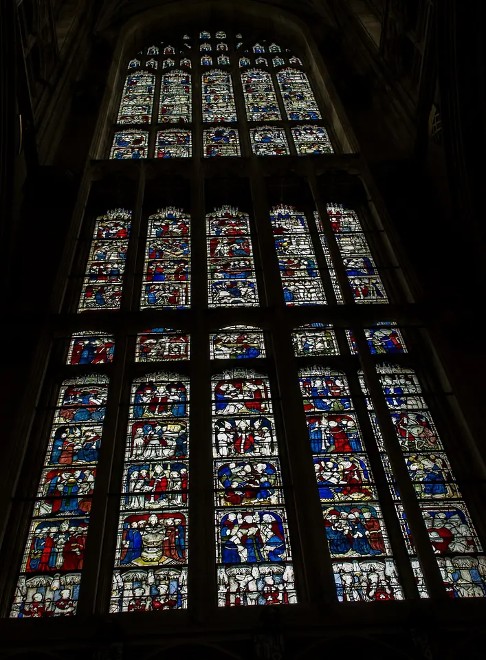 A towering medieval stained glass window divided into dozens of small rectangular panels of red, blue and white glass, each showing a scene from a saint's life