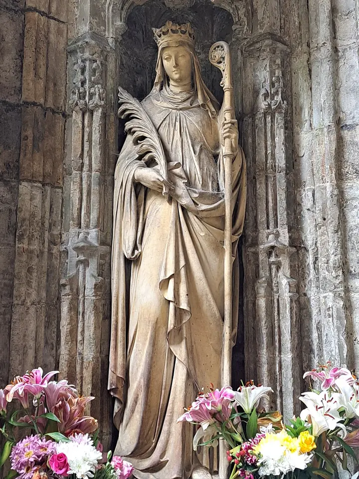 Stone statue of a crowned woman holding a palm and an abbess's staff in a Gothic niche, with lilies and flowers at her feet
