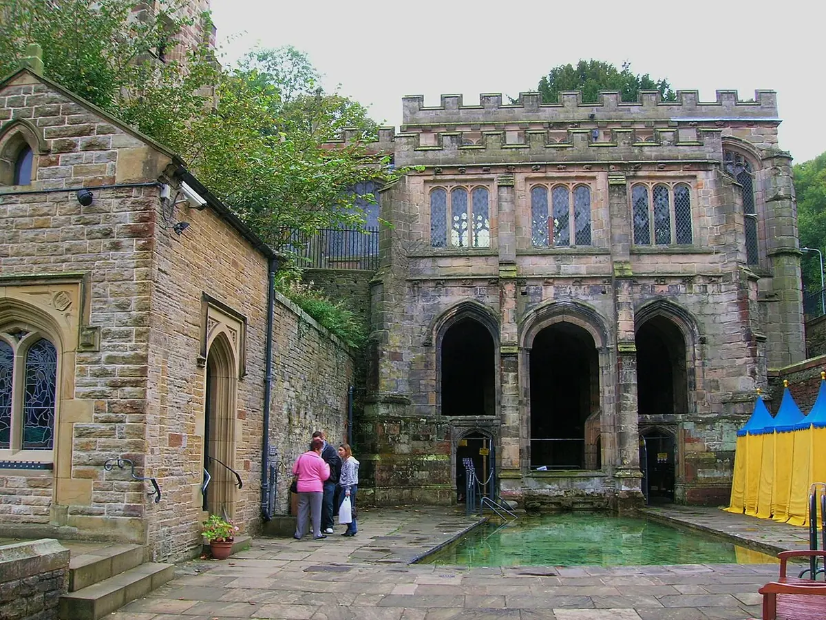A stone Gothic well house with three arches and battlements beside an outdoor bathing pool of green water, with yellow and blue changing tents