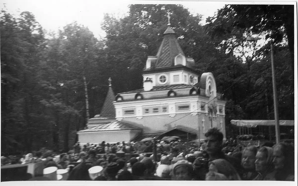 Black-and-white photograph of a large crowd of people gathered in front of a small chapel with a tent roof and a cross among trees