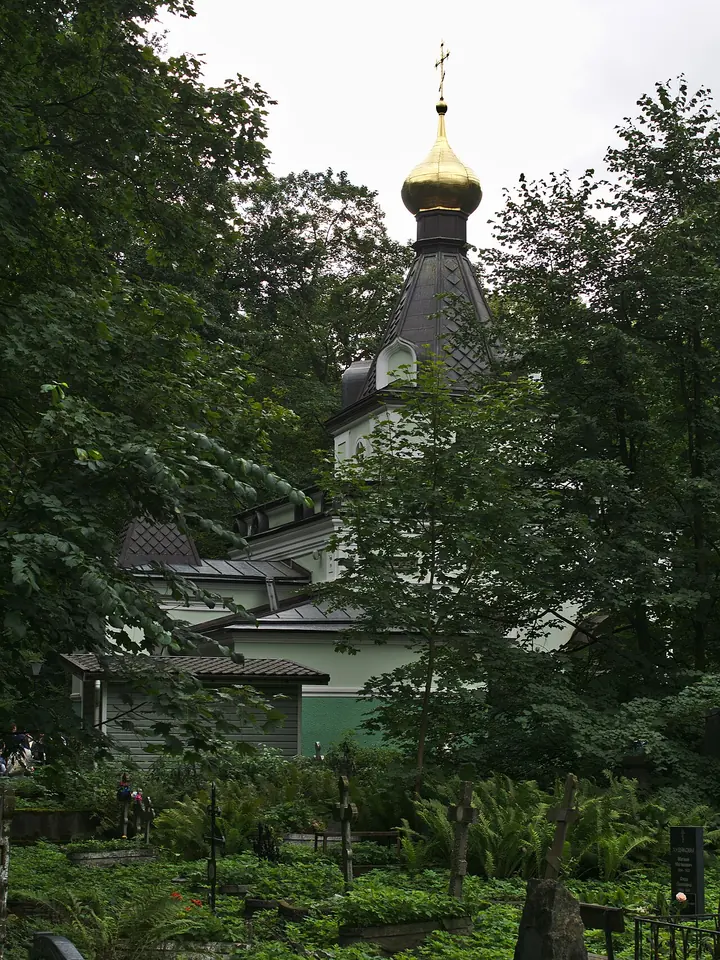 A small pale green chapel with a pointed tent roof and a gold onion dome among tall trees and cemetery crosses