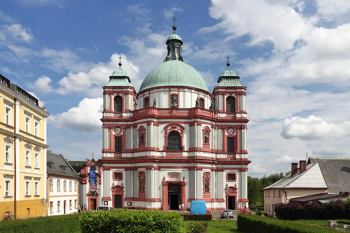 A large pink and white Baroque church with a green dome and two towers under a blue sky with clouds