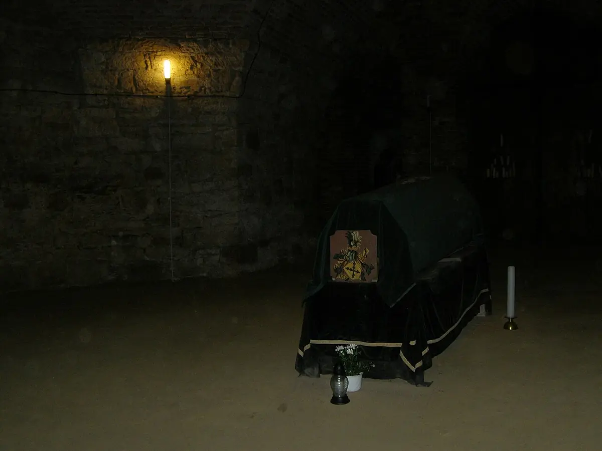 A dark vaulted crypt lit by a single lamp, with a coffin draped in black standing in the middle