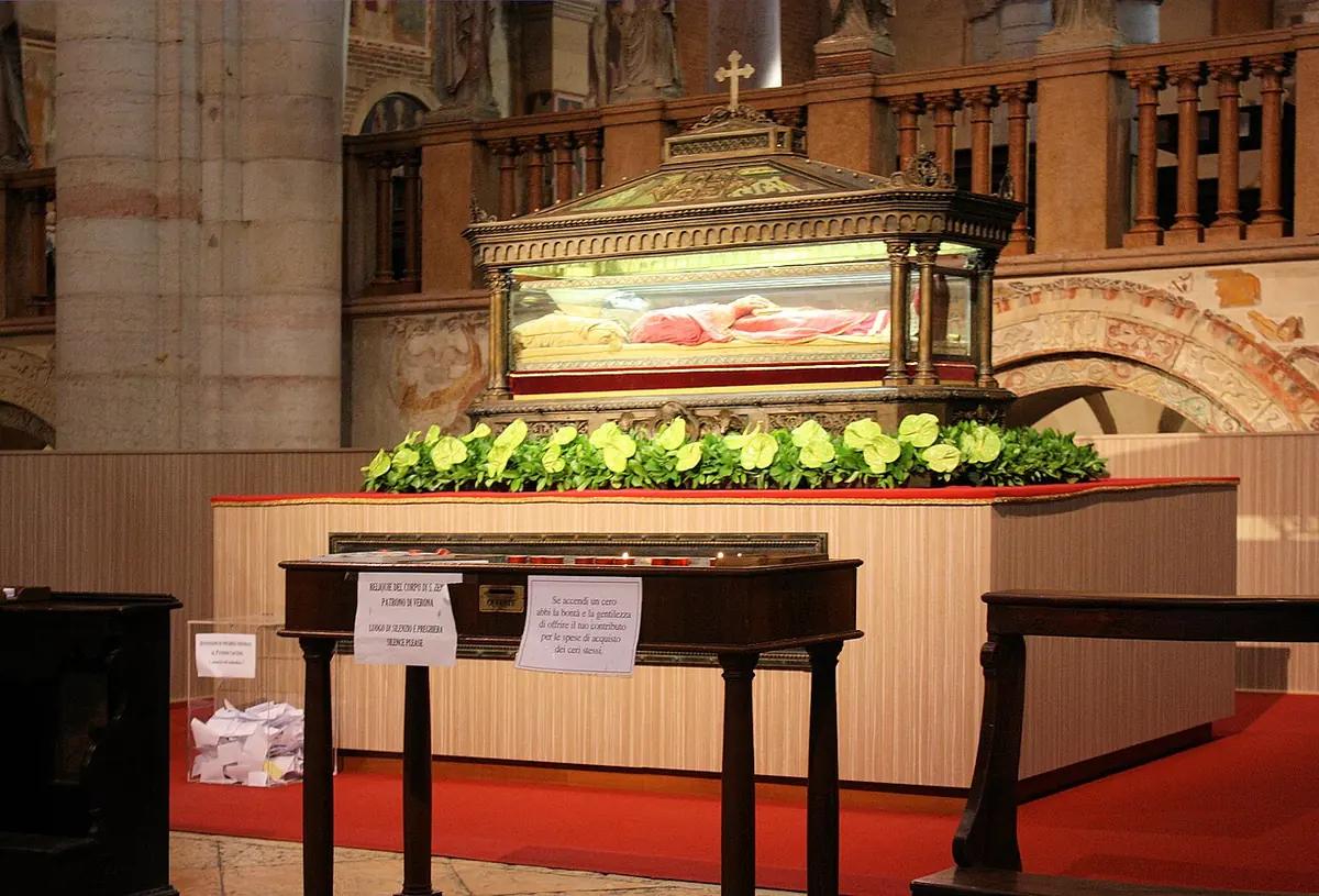 A glass-sided reliquary urn on a draped stand in a church crypt, holding the vested figure of a bishop, with flowers along its base