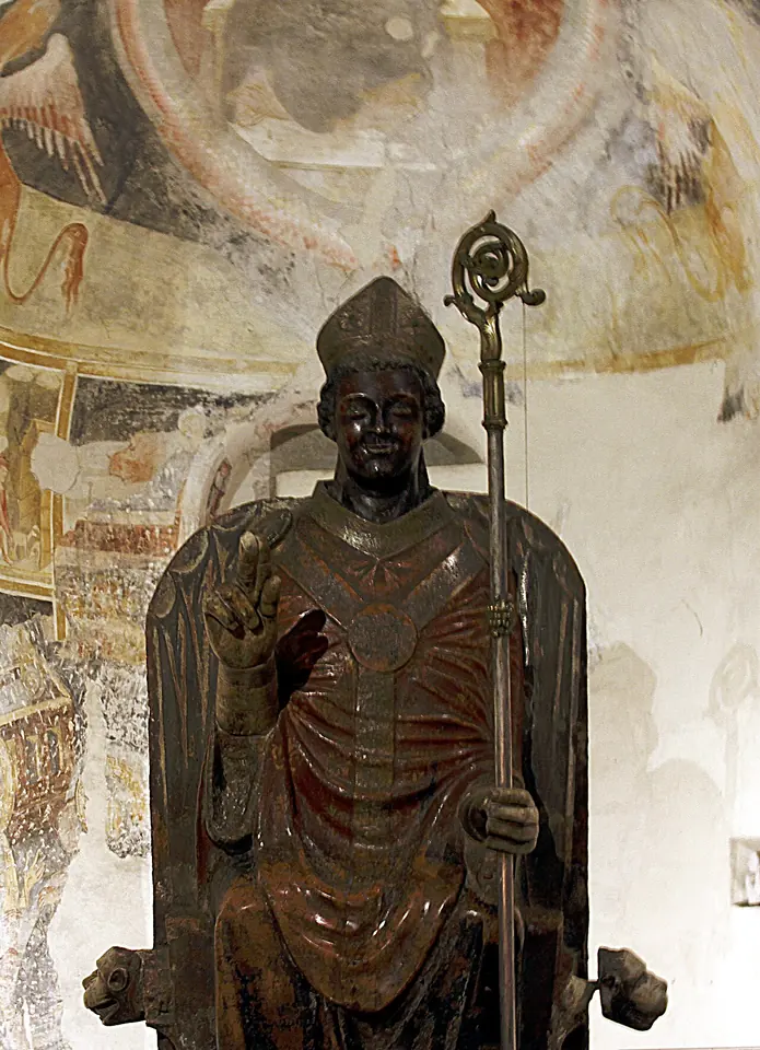 Painted marble statue of a dark-faced seated bishop in a tall miter, raising his right hand in blessing and holding a crozier, in front of faded medieval frescoes