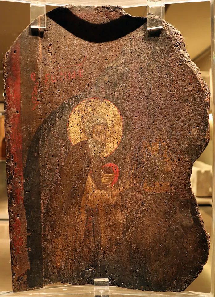 Worn, damaged medieval icon panel showing a haloed monk in dark robes holding a red chalice