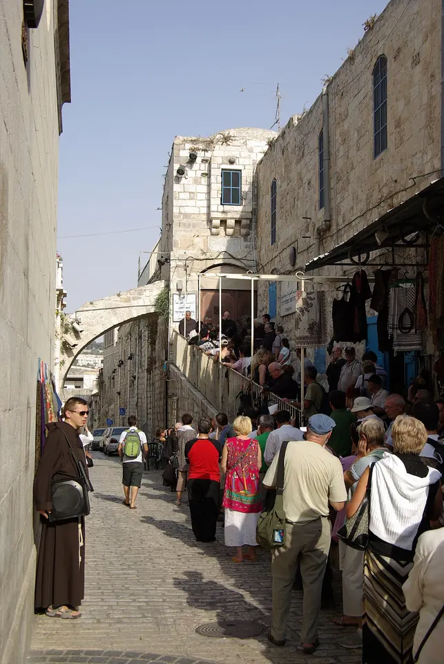 Peregrinos y turistas llenan una calle estrecha de piedra de la Ciudad Vieja de Jerusalén bajo un arco, con un fraile franciscano de hábito pardo a la izquierda