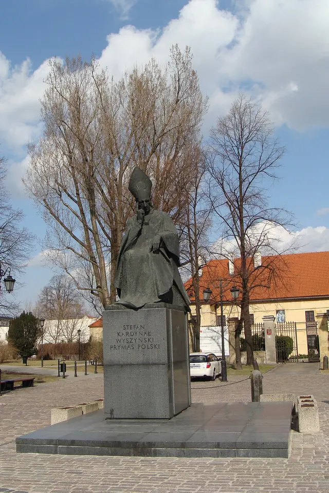 A dark bronze statue of a seated cardinal on a tall granite pedestal in a paved square with bare trees and a red-roofed building behind