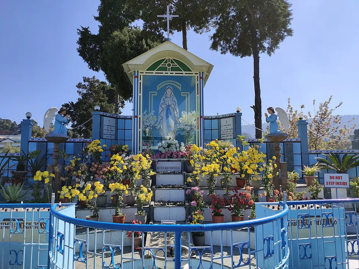 A small outdoor shrine with an image of Mary under a pointed roof, surrounded by yellow flowers, blue railings and tall pine trees