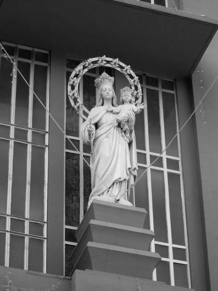Black-and-white photograph of a white statue of the crowned Virgin holding the Child Jesus, framed by a halo of stars in front of a tall window