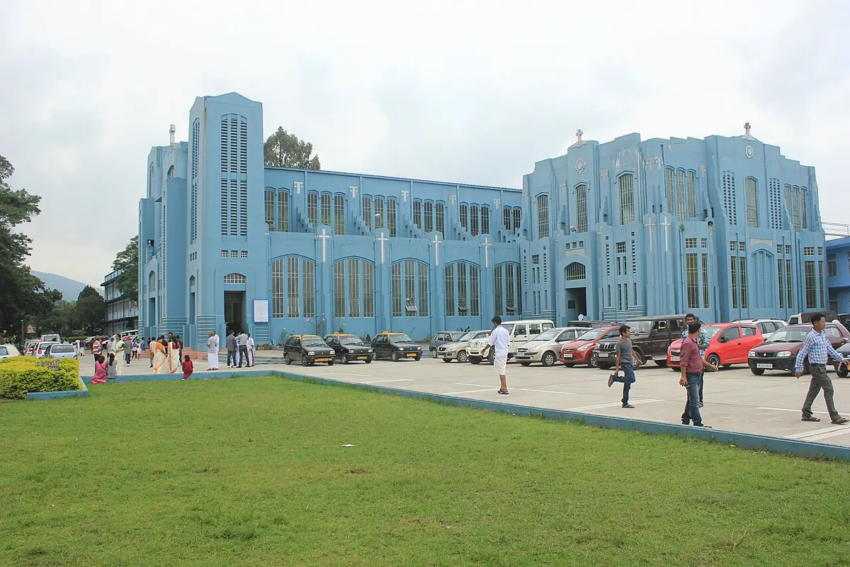 The long, pale blue facade of Shillong's cathedral with tall vertical towers and a cross on top, seen across a lawn with people walking in front