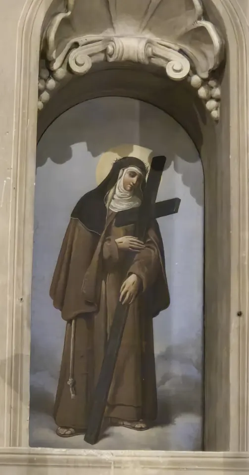 Painting in a carved niche of a Capuchin nun in a brown habit and black veil, wearing a crown of thorns and embracing a large wooden cross