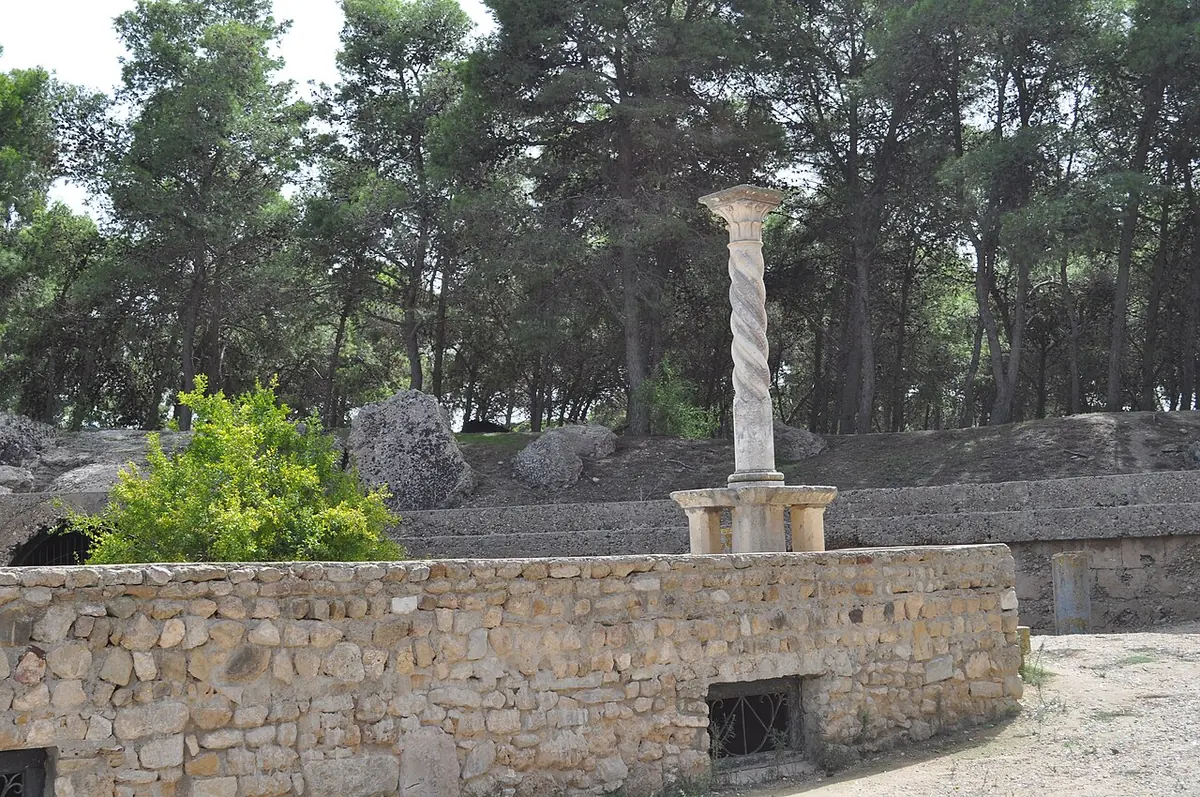 A low curved stone wall in the ruined arena of Carthage under pine trees, with a single tall column with a spiral shaft standing in the middle