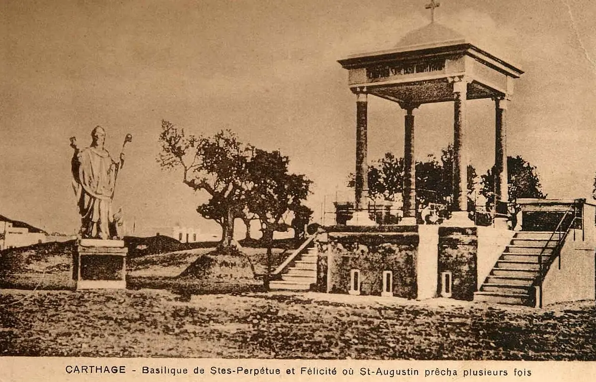 Old sepia postcard of a small domed canopy on columns, reached by a stair, with a statue of a bishop beside it, on the bare ground of the Basilica Majorum at Carthage
