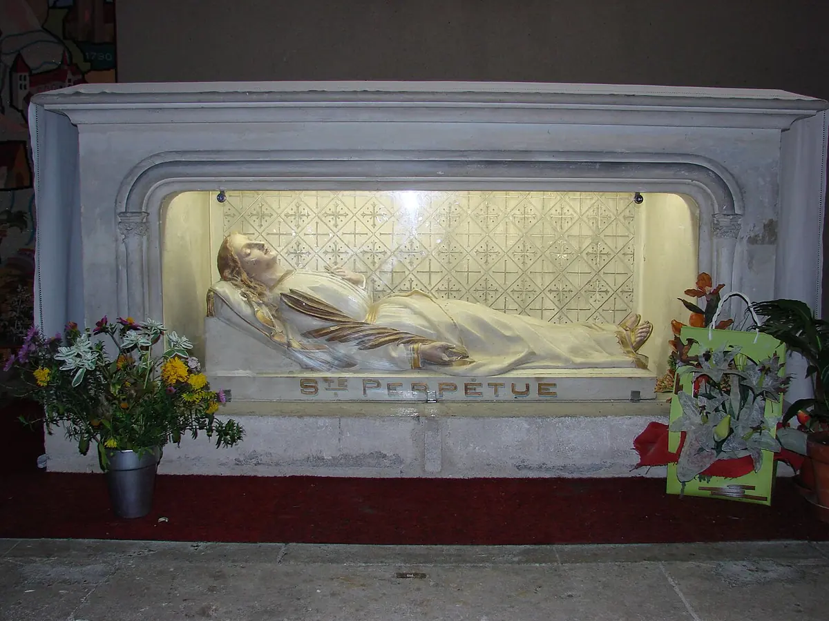 A white marble shrine with a recumbent figure of St. Perpetua behind glass, lettered Ste Perpétue, with flowers placed in front of it