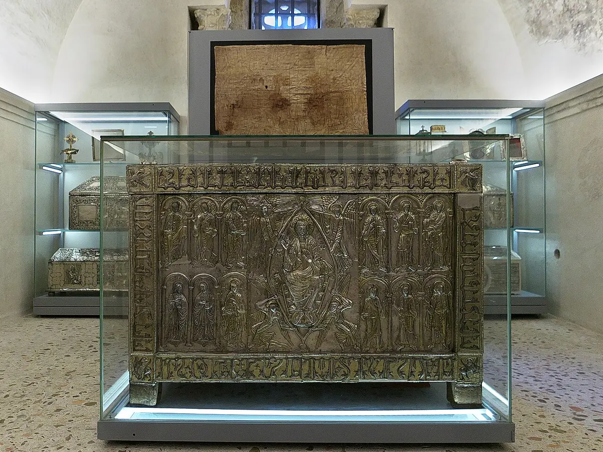 A large chest covered with silver plates showing Christ in a mandorla surrounded by apostles, in a glass case in a white chapel, with a framed cloth hanging on the wall behind