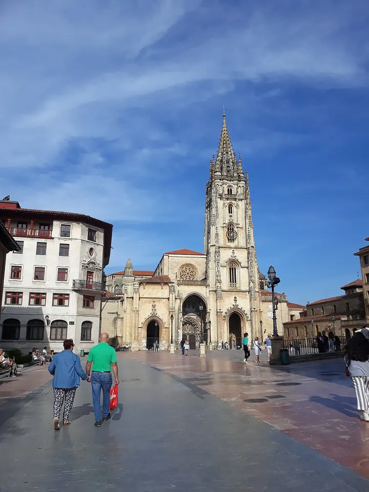 A Gothic cathedral with a tall single spire at the end of a broad paved square under a blue sky, with people walking toward it