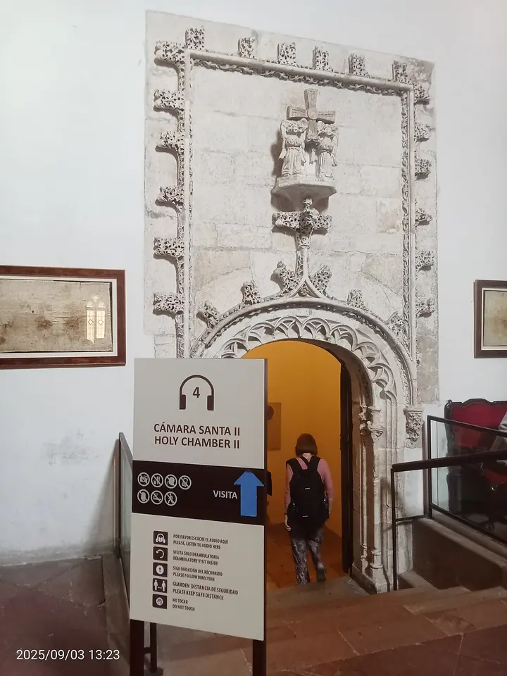 A carved Gothic doorway with a small statue above it, opening onto steps that lead up into the Cámara Santa, with a visitor going in