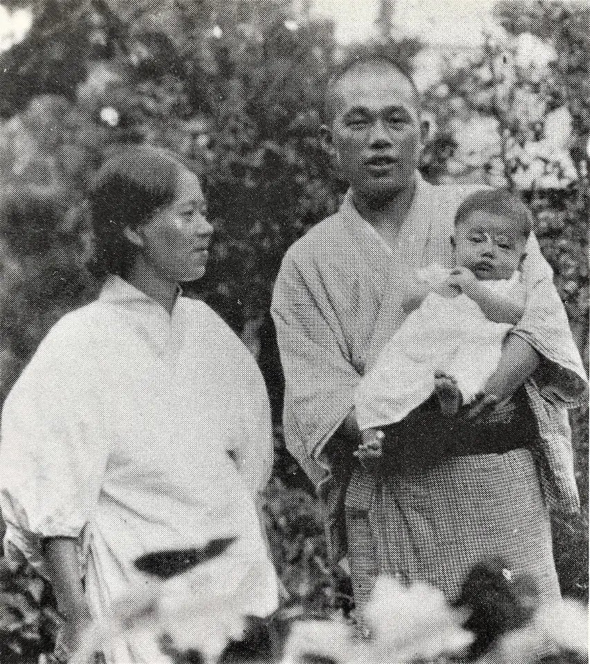Black-and-white photograph of a smiling young couple in light summer kimonos outdoors, the father holding their baby son