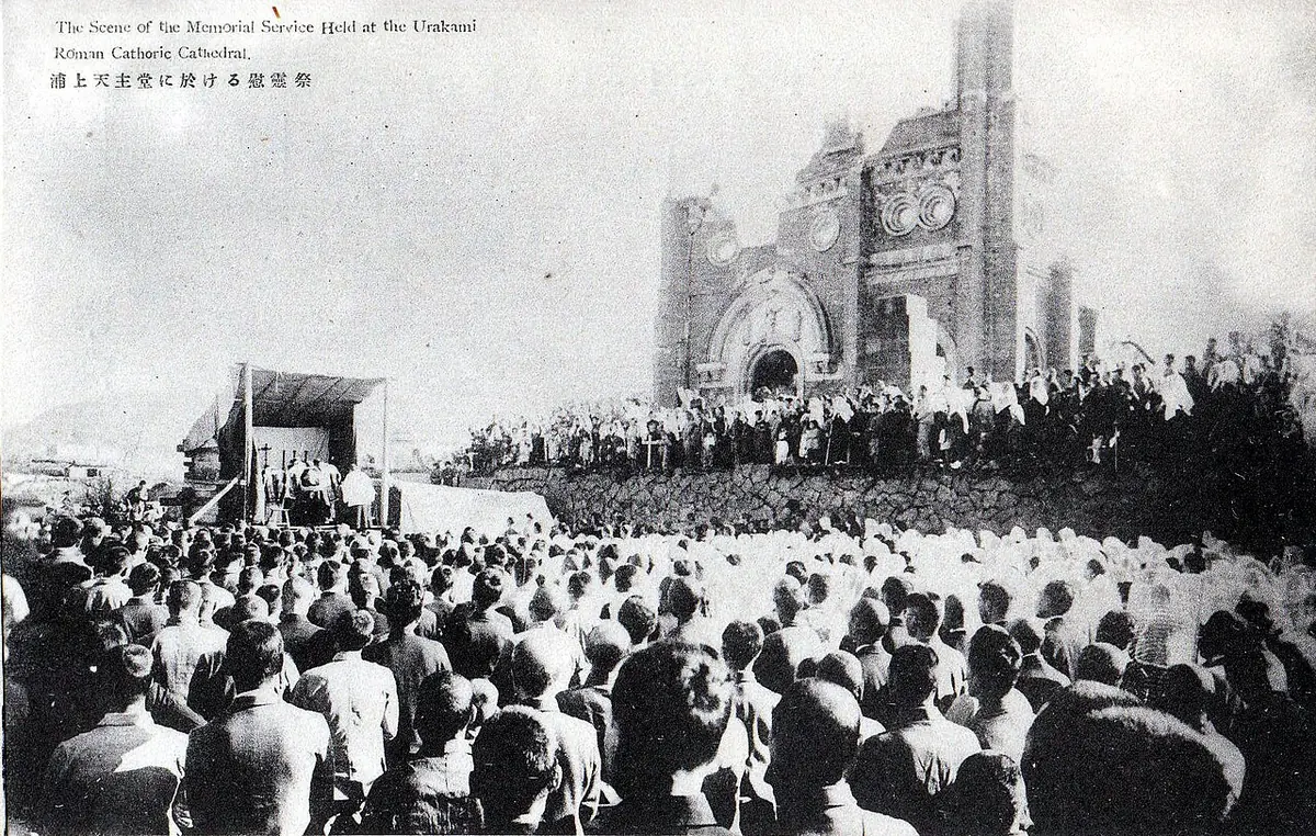 Old postcard photograph of a vast crowd gathered on open ground before the gutted walls and tower of Urakami Cathedral, facing a small covered altar