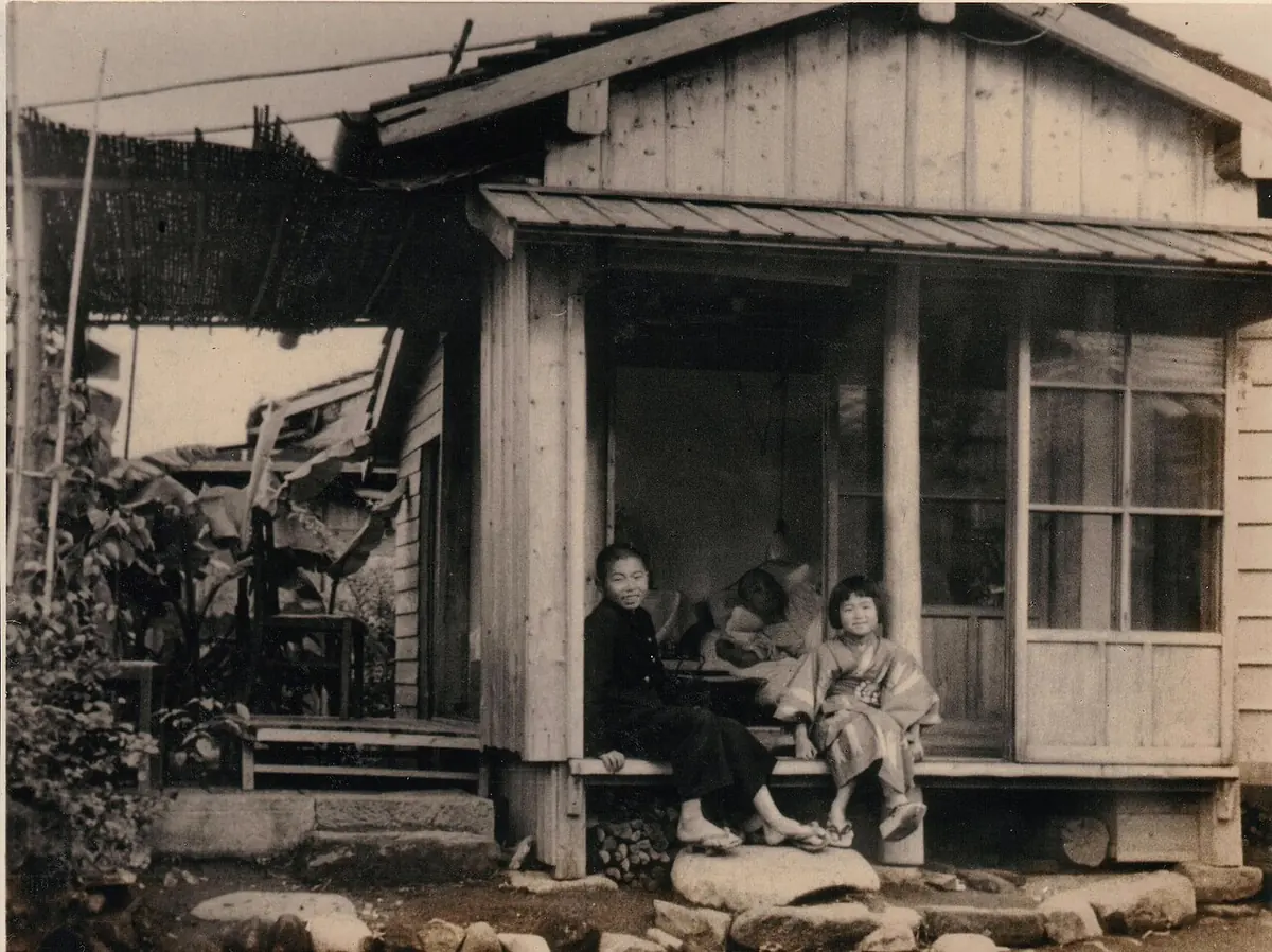 Old photograph of a tiny wooden hut with a porch, where a man lies inside while two children sit on the step outside