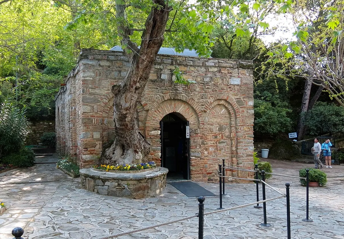 A small stone chapel with an arched doorway and a large tree in front, on a paved terrace among green trees
