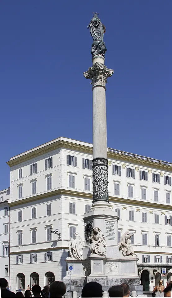 A tall marble column topped by a bronze statue of the Virgin, rising from a sculpted base in front of a pale city building