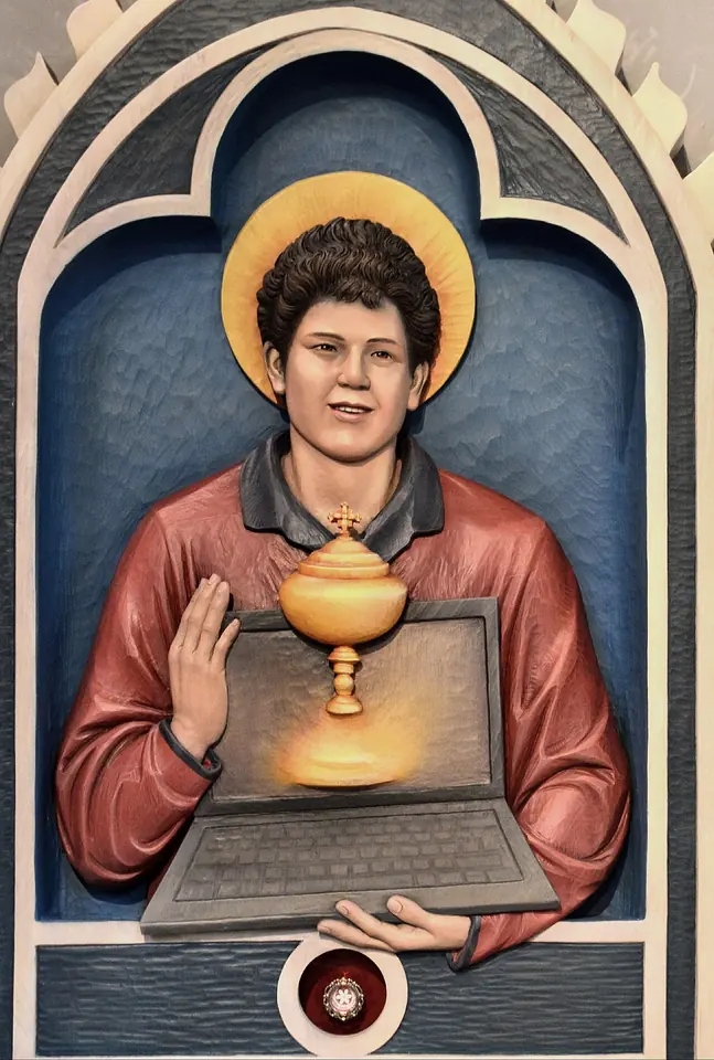 Painted carved relief of a smiling curly-haired teenager with a gold halo in a red sweater, holding an open laptop with a golden chalice and Host rising from it