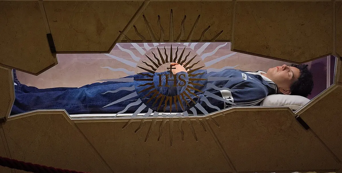 A glass-fronted tomb in which the body of a teenager in jeans, sneakers and a sweatshirt lies on a white pillow, behind a stone slab carved with a gold sunburst and the letters IHS