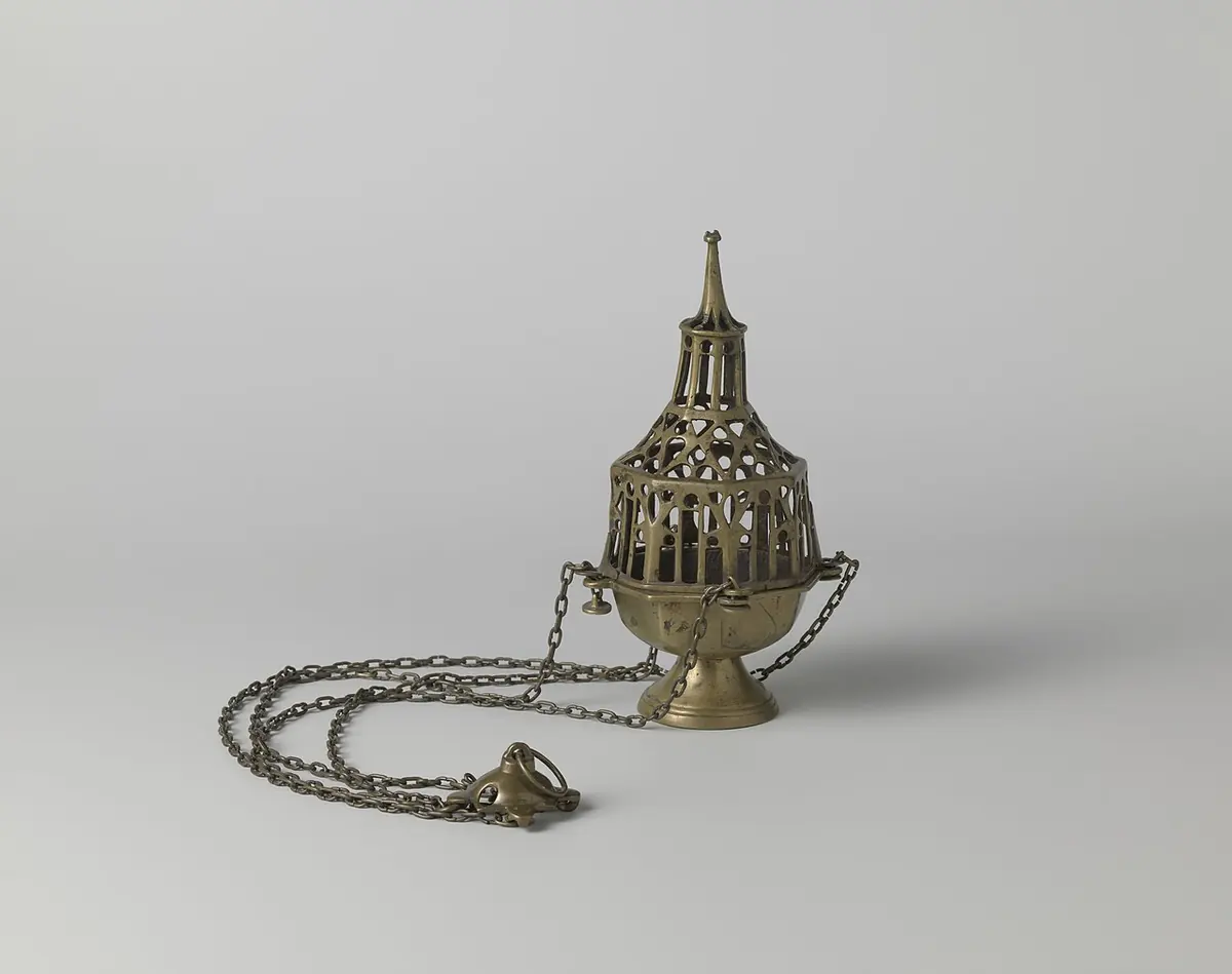 A late medieval cast-metal thurible with a round bowl on a footed base and a tall, pierced, spire-shaped lid like a Gothic tower, its chains coiled beside it