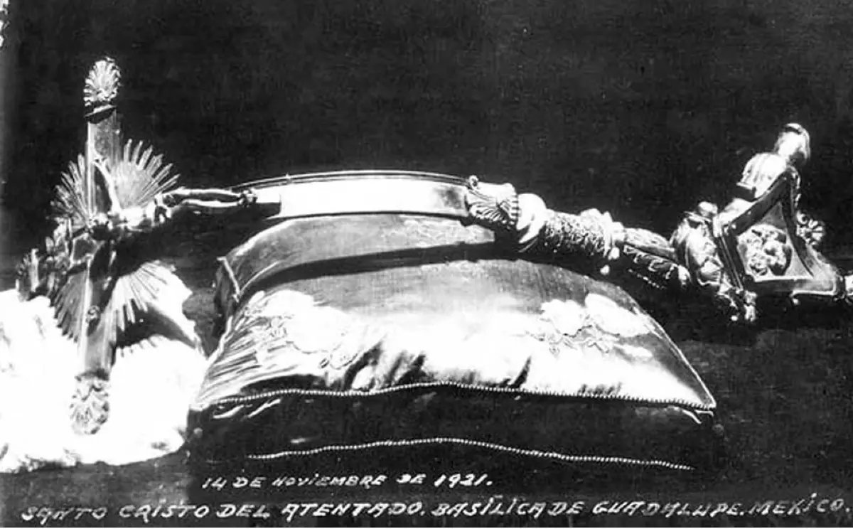 Old black-and-white photograph of a metal crucifix bent backward lying on a cushion, with a handwritten caption dated November 14, 1921