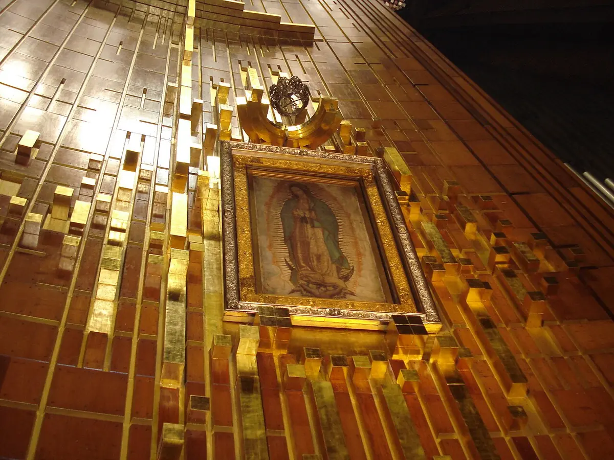 The framed image of Our Lady of Guadalupe hanging on a wall of gold-colored panels in the basilica, seen from below at an angle