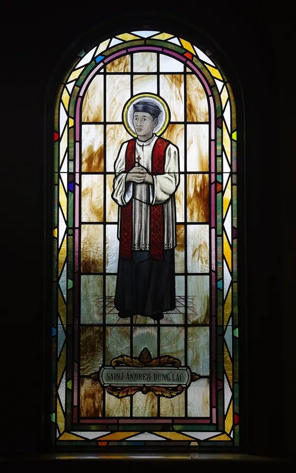 Arched stained-glass window of a Vietnamese priest in black cassock, white surplice and red stole, standing with hands joined, his name on a panel at his feet