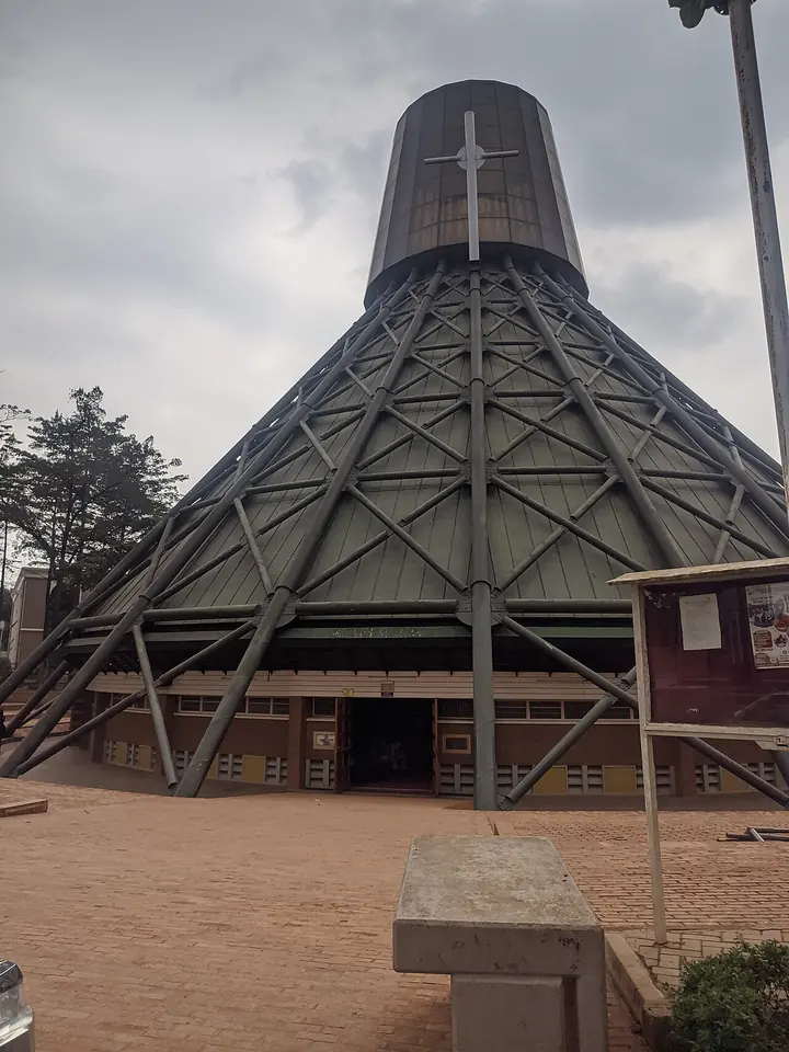 Tall cone-shaped basilica of steel and concrete with a cross at its peak, standing over a red-earth courtyard under a gray sky