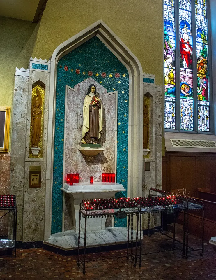 Side shrine with a white statue of St. Thérèse in a niche of green mosaic, with rows of red votive candles in front and a stained-glass window beside it