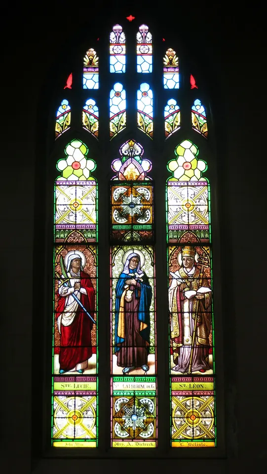 A tall Gothic stained-glass window with three standing saints in colored robes, a woman in the center between a young woman and a pope