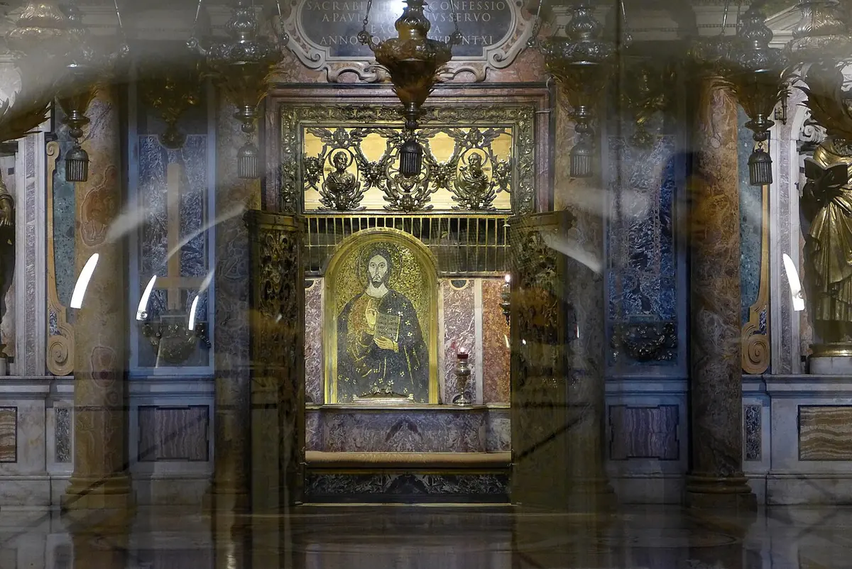 A gilded niche with a small mosaic of Christ, framed by marble and bronze, seen through glass in the lower level of a basilica