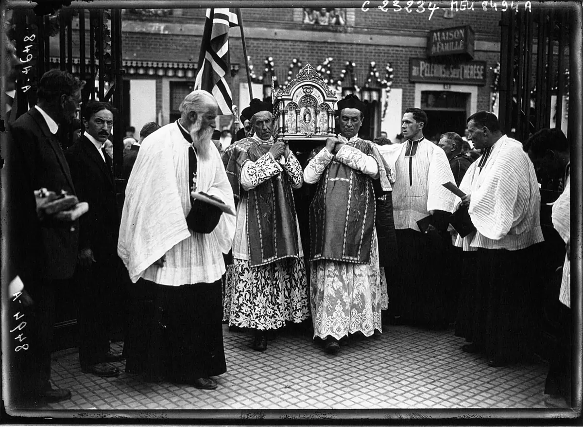 Black-and-white press photograph of priests in vestments and surplices carrying a small ornate reliquary through a crowded doorway