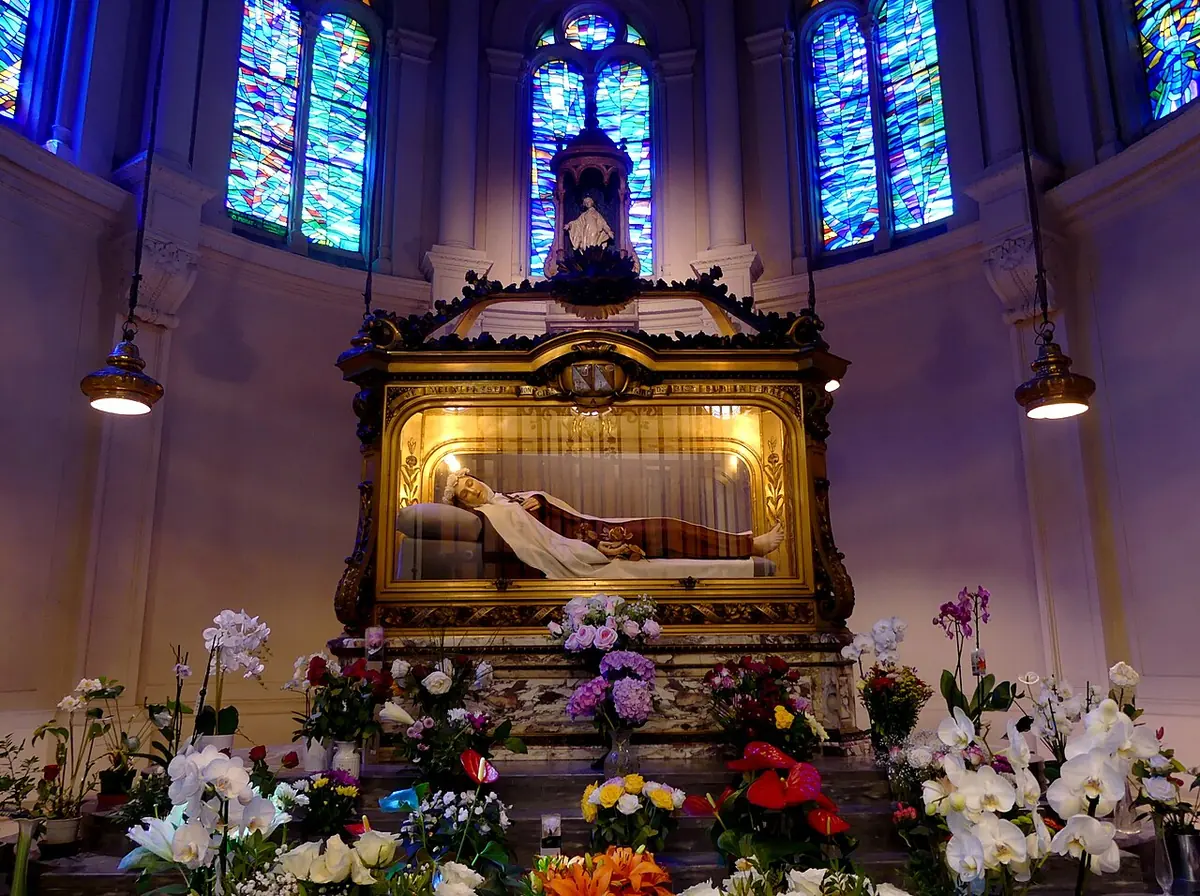A gilded glass-sided shrine holding a reclining figure of a Carmelite nun, surrounded by bouquets of flowers under blue stained-glass windows