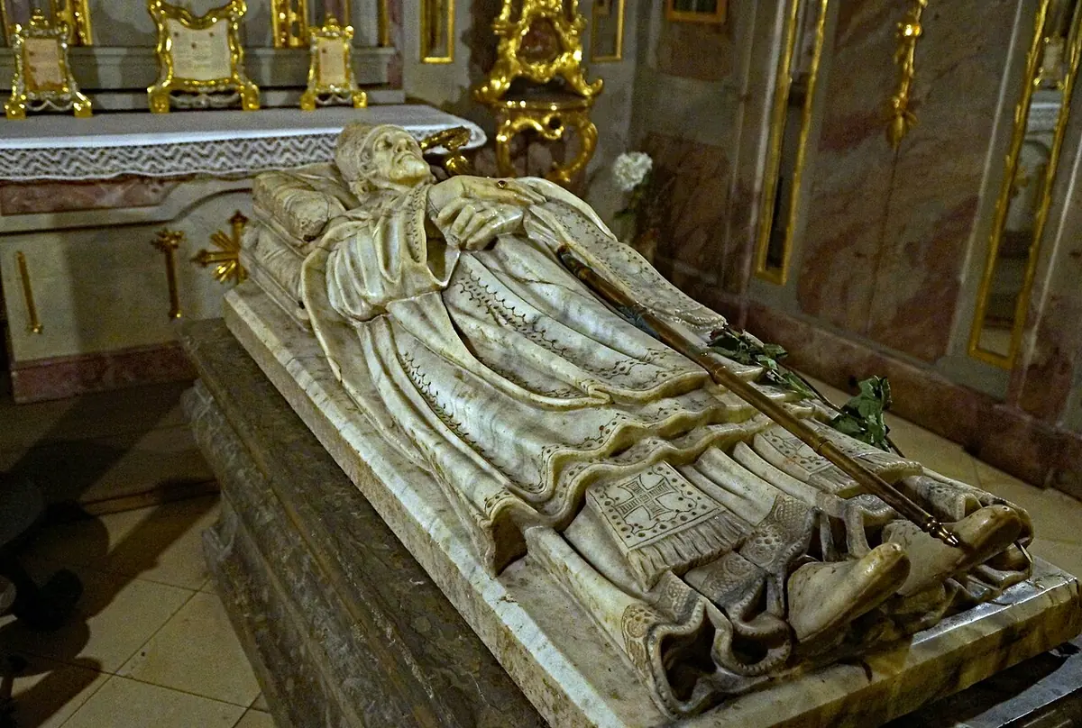 Pale stone tomb with a carved figure of a bishop lying on the lid, in a crypt with gilded decoration behind it