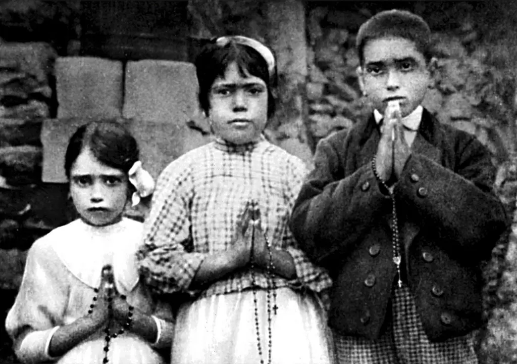 Black-and-white photograph of two small girls and a boy standing against a stone wall, holding rosaries and joining their hands in prayer