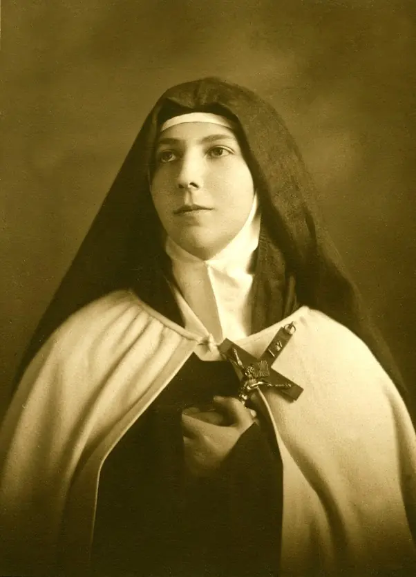 Sepia photograph of a young Carmelite nun in a dark veil and white mantle, holding a crucifix to her chest and looking past the camera